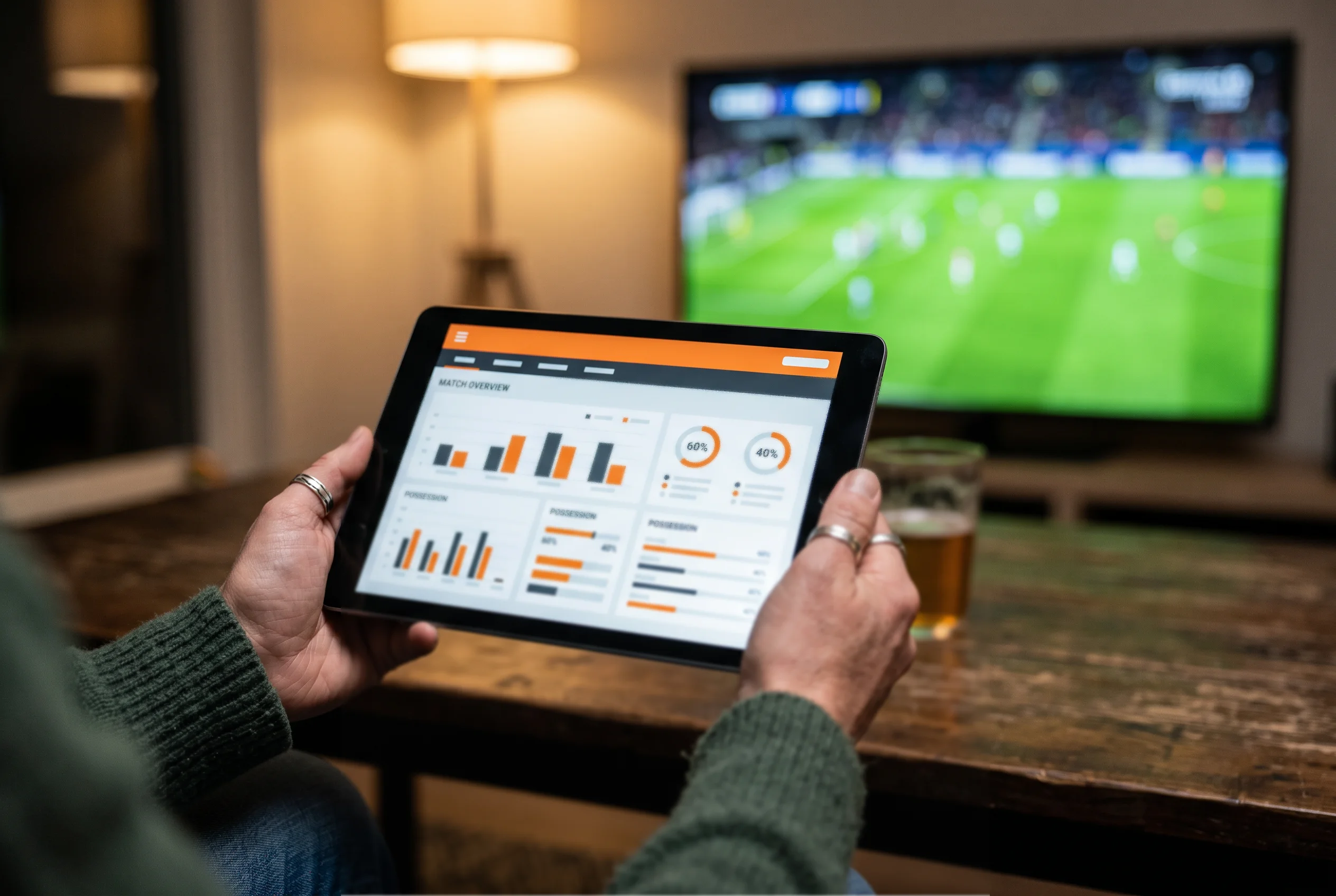 Persoon analyseert wedstrijdstatistieken op een tablet met een Eredivisie-wedstrijd op de achtergrond